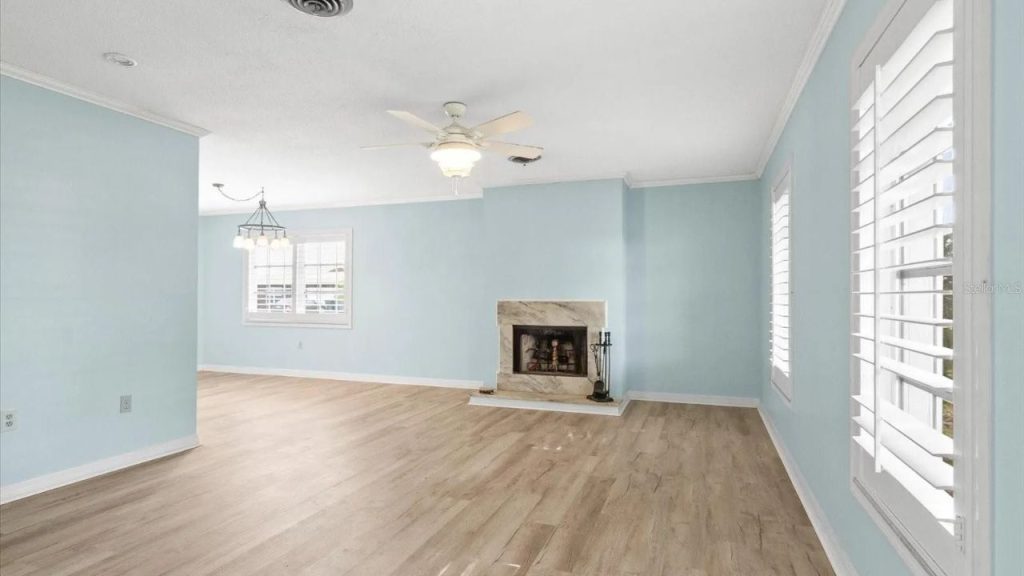 A bright living room with pastel blue walls, light wood floors, a marble fireplace, and large windows with white shutters, lit by a ceiling fan with a light.
