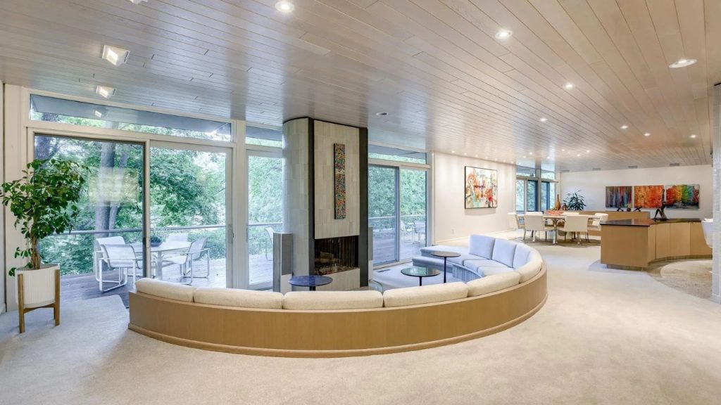 Curved living room with fireplace and forest views through tall windows.