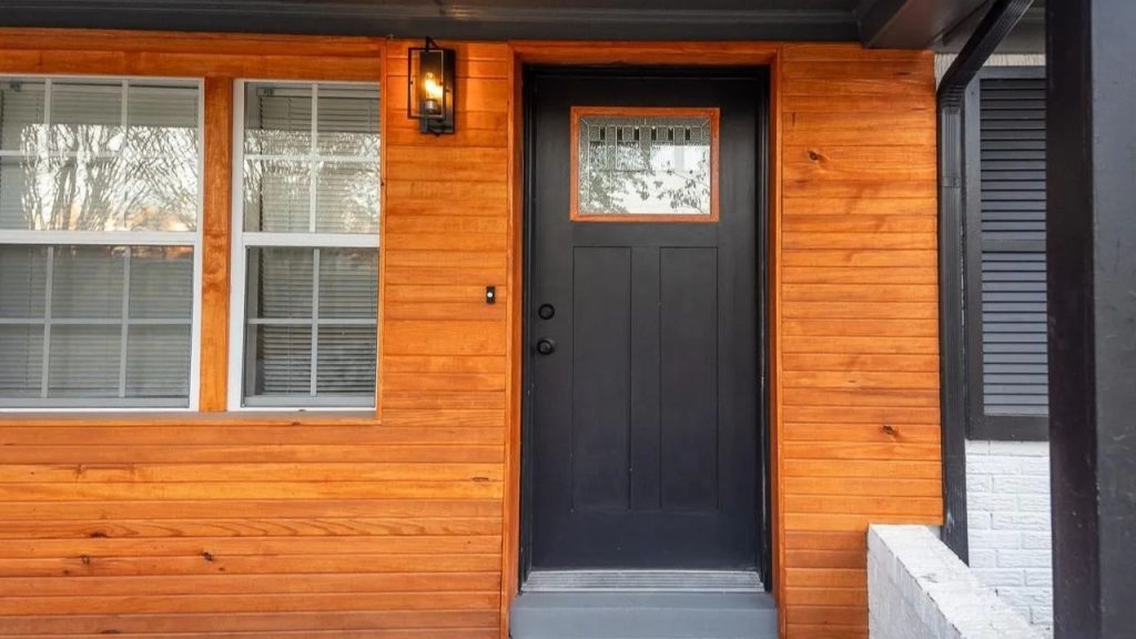 Black front door with glass panel set in warm wood siding, flanked by windows.