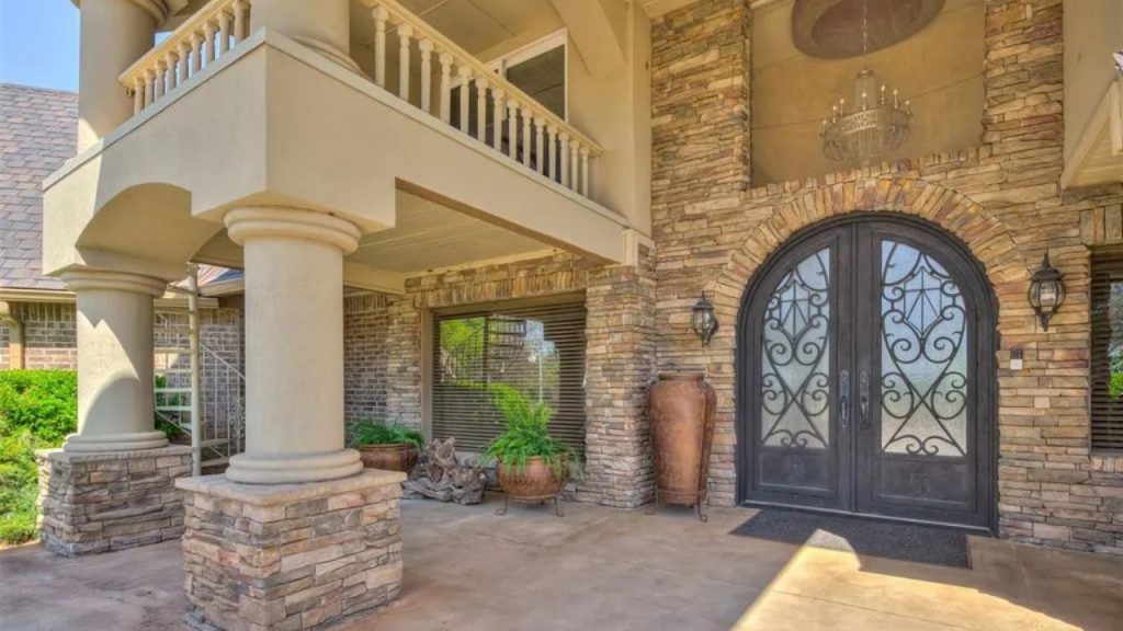 Elegant entrance with ornate double doors, stone columns, and a chandelier, surrounded by greenery and pottery.