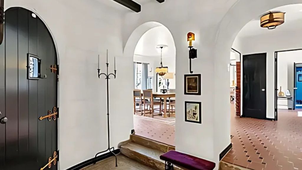 A stylish hallway with arched doorways, dark wooden doors, terracotta tiles, and a cozy bench, leading to a warmly lit dining area.