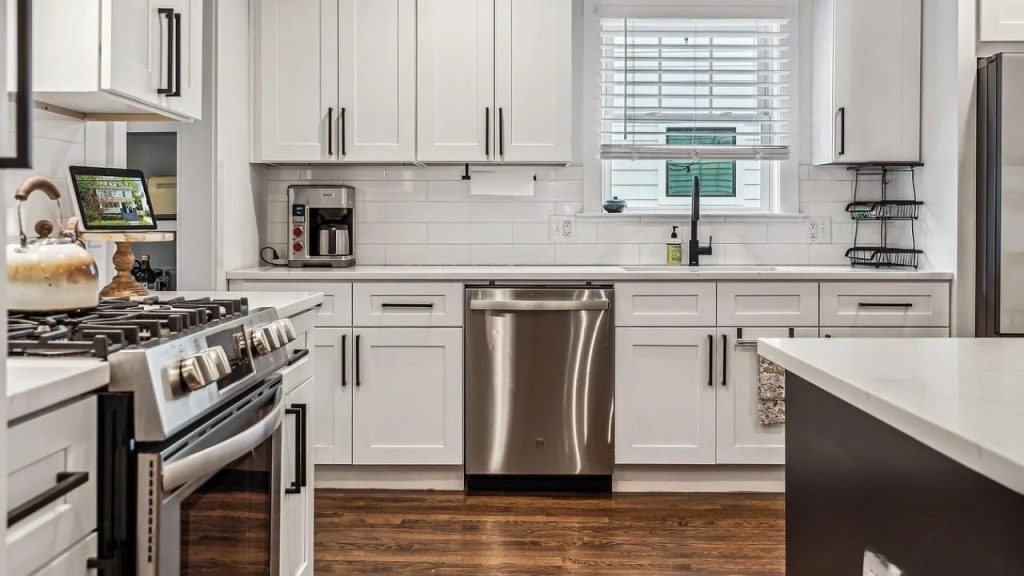 A bright kitchen with white cabinets, stainless steel appliances, and large windows with shutters.