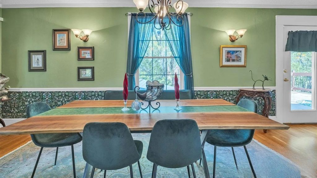 A stylish dining area with a wooden table, teal velvet chairs, green walls, a chandelier, and a window with blue curtains.
