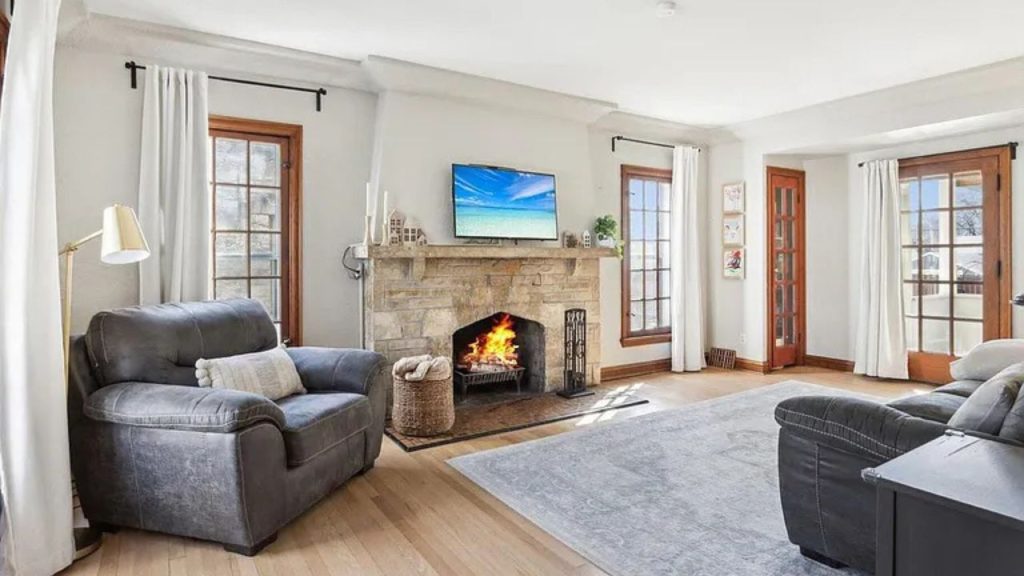 A cozy living room with a stone fireplace, gray armchairs, and large windows with white curtains.