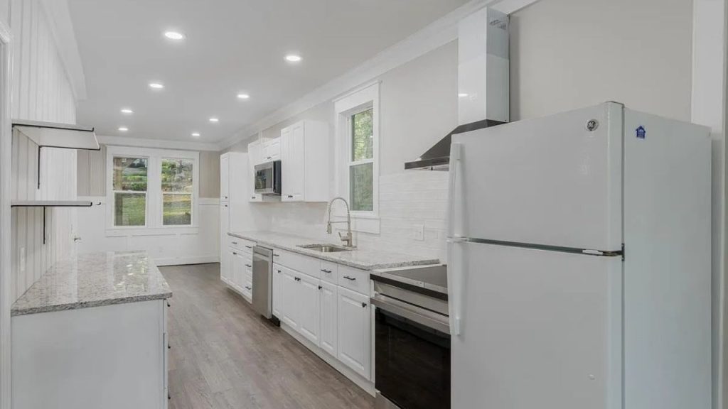 A bright kitchen with white cabinets, granite countertops, modern appliances, and large windows overlooking greenery.