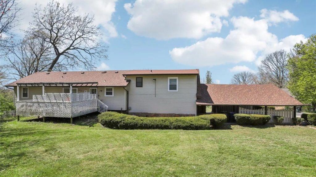 A cozy backyard with a spacious deck, light-colored house, and lush green lawn, framed by trees and bushes under a bright, cloudy sky.