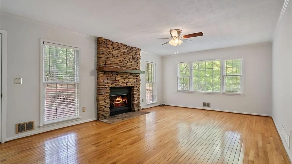 A cozy living room with a stone fireplace, wooden floors, large windows, and a ceiling fan, bathed in natural light.
