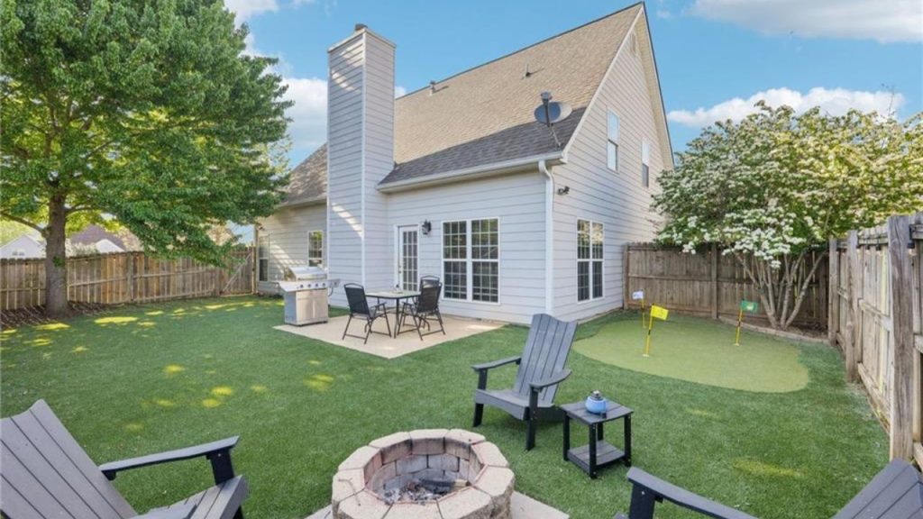 A cozy backyard with a fire pit, chairs, a putting green, and a grill area, framed by trees and a white house.