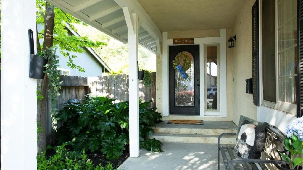 A cozy front porch with a bench, hydrangeas, hanging plants, and a decorative door, framed by white pillars and greenery.