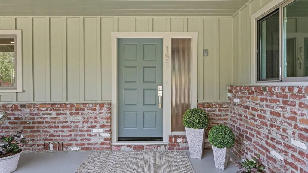 A stylish front entrance with a green door, brick base, and potted topiaries, framed by large windows.