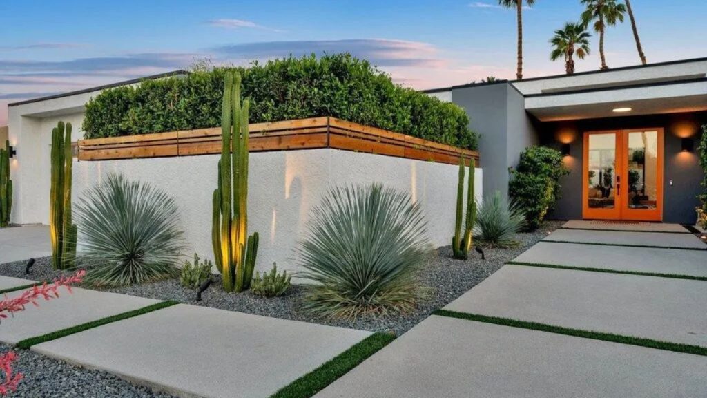 A stylish desert modern entrance with an orange door, cacti, and a green planter box under a twilight sky.