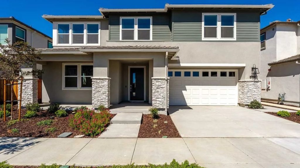 A modern two-story suburban home with a gray and teal exterior, stone pillars at the entrance, a double garage, and a tidy front yard with young trees under a clear blue sky.