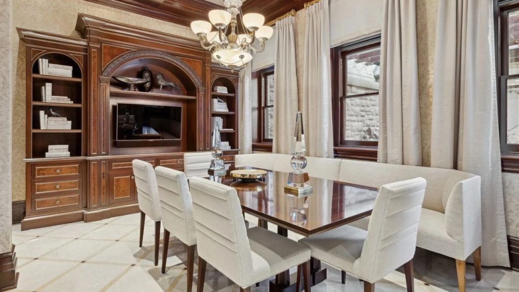 A refined dining room with a wooden table, white chairs, a built-in bench, and mahogany cabinetry, lit by a chandelier and framed by cream curtains.