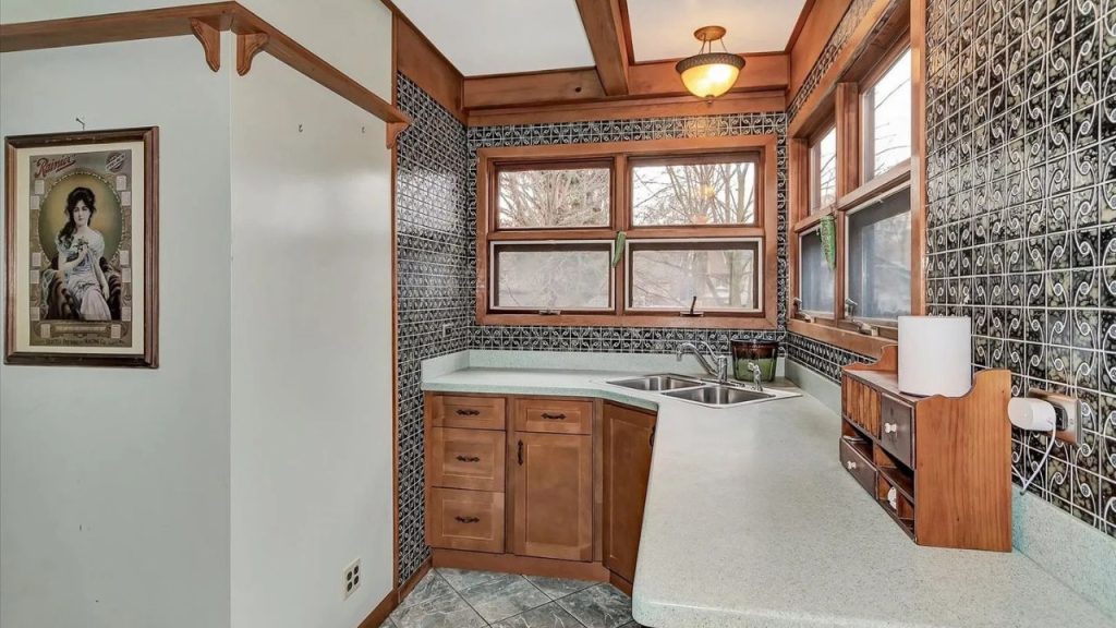 A vintage kitchen with black-and-white tiled walls, wooden cabinets, exposed beams, a double sink, and a classic portrait, lit by a warm pendant light.