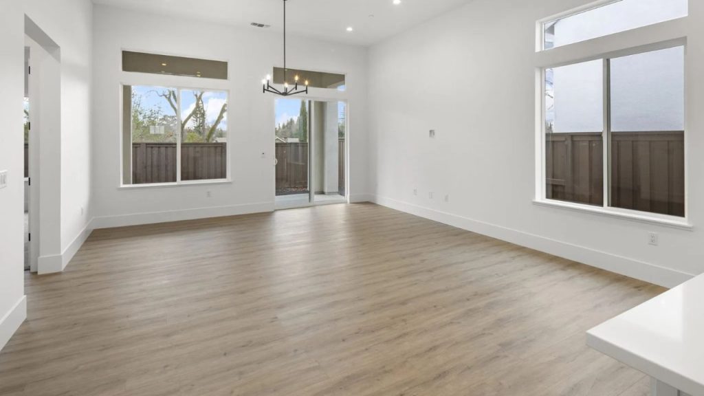 A spacious great room with light wood floors, white walls, large windows, a sliding glass door to a patio, and a modern chandelier.
