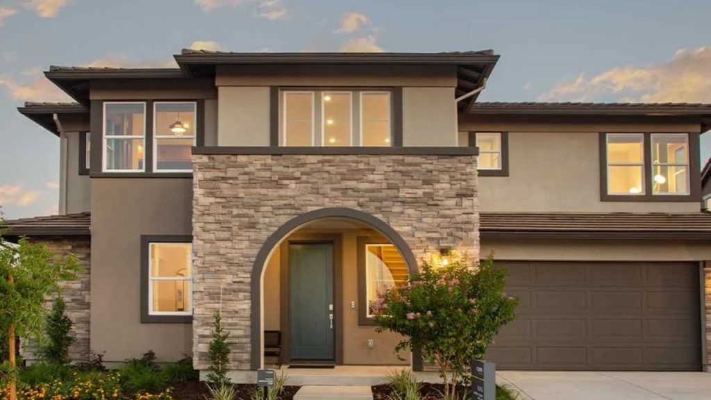 A two-story home with a stone-accented entrance, large windows, and a warm glow from interior lights at dusk.
