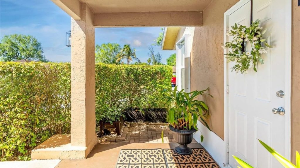 A cozy porch with a white door, floral wreath, potted plant, and lush hedges under a blue sky.