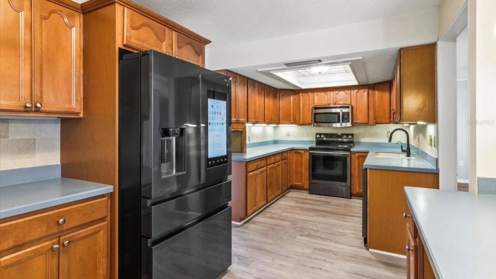 A modern kitchen with warm wooden cabinets, black appliances, a smart fridge, gray countertops, and light wood floors, softly lit by under-cabinet lights.