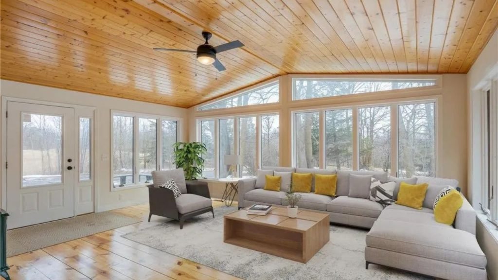 Sunroom with pine ceiling, large windows, gray sectional, and forest view.