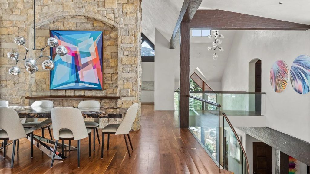 A modern dining area with a stone wall, abstract art, and a chic chandelier, overlooking a glass-railed mezzanine with colorful wall pieces.