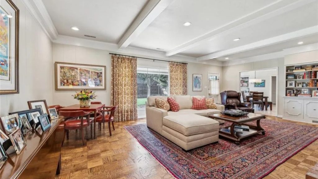 A cozy living room with a sectional sofa, colorful rug, dining area, and large windows, featuring hardwood floors and bookshelves.