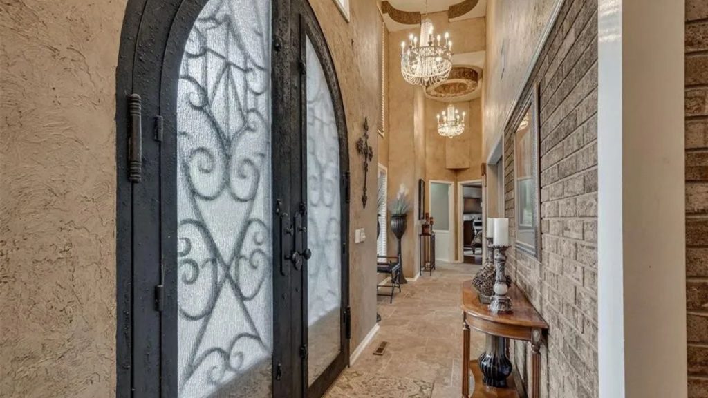 Spacious foyer with ornate glass doors, chandeliers, and a console table, framed by brick walls and high ceilings.