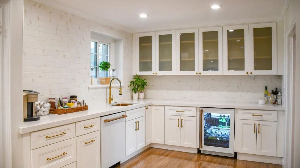 A sleek kitchen with white cabinets, gold accents, a wine cooler, and a subway tile backsplash, accented with plants.