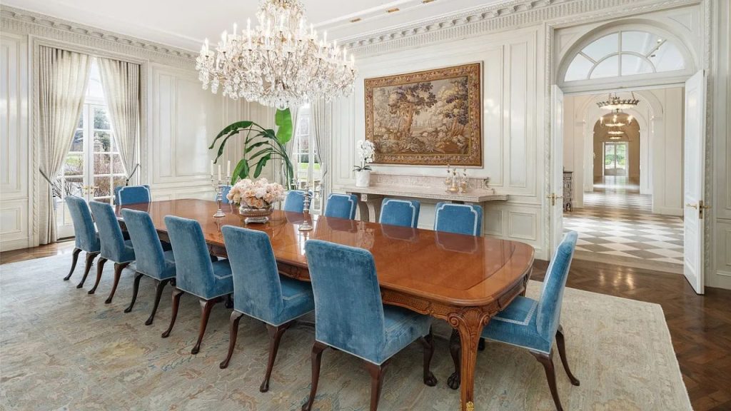 A grand dining room with a long table, blue chairs, a chandelier, and large windows.