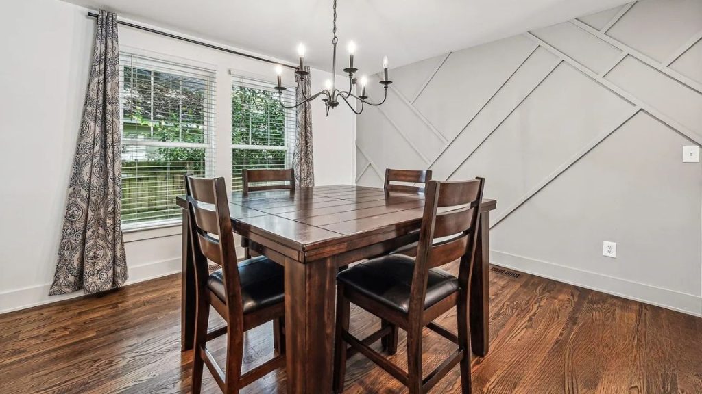 A dining area with a wooden table, chandelier, and geometric wall design by large windows.