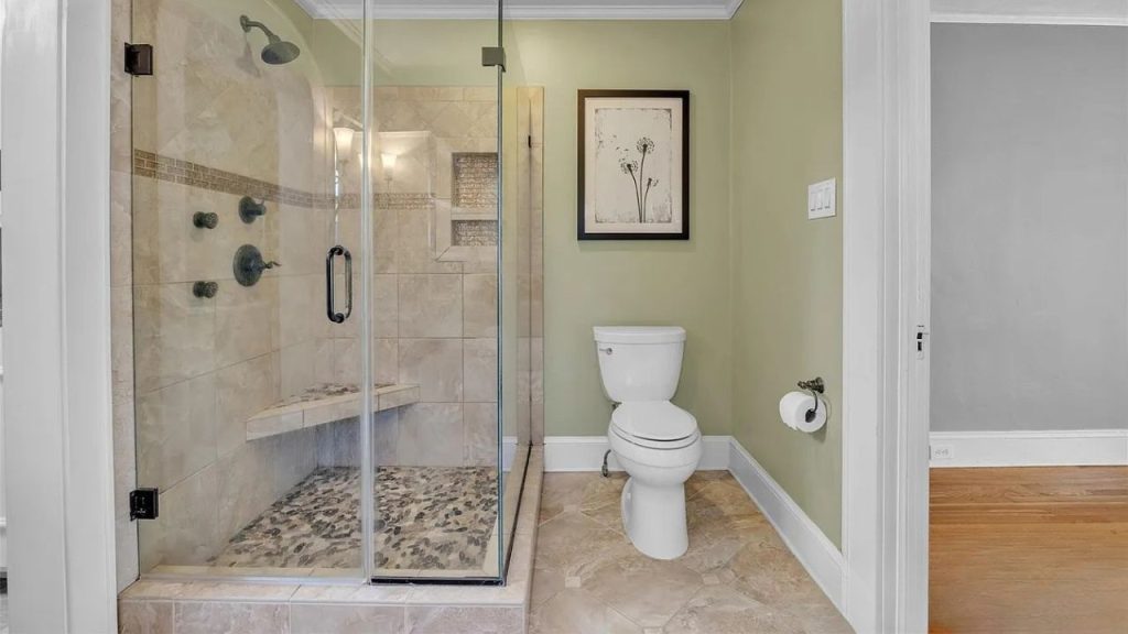 A serene bathroom with a glass shower, pebble floor, green walls, white toilet, and framed floral art.