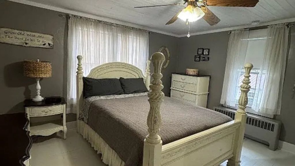 A serene bedroom with a four-poster bed, gray quilt, rustic nightstand, and large windows with sheer curtains.