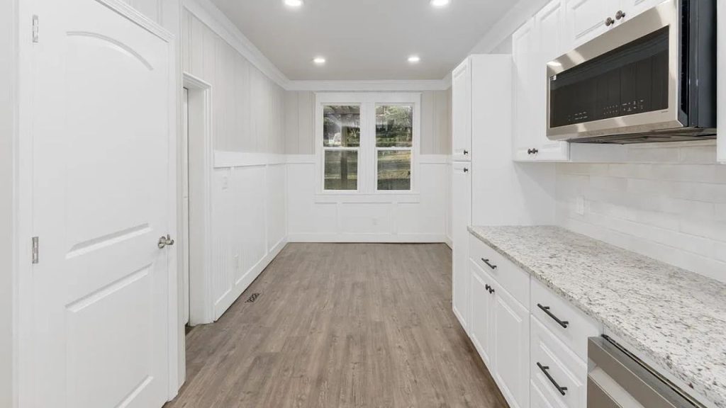 A cozy kitchen with white cabinets, granite countertops, stainless steel appliances, and large windows with greenery views.