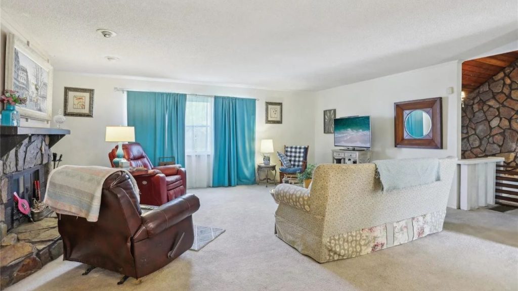 A cozy living room with a red recliner, floral sofa, stone fireplace, and blue curtains, adorned with art and a round mirror.