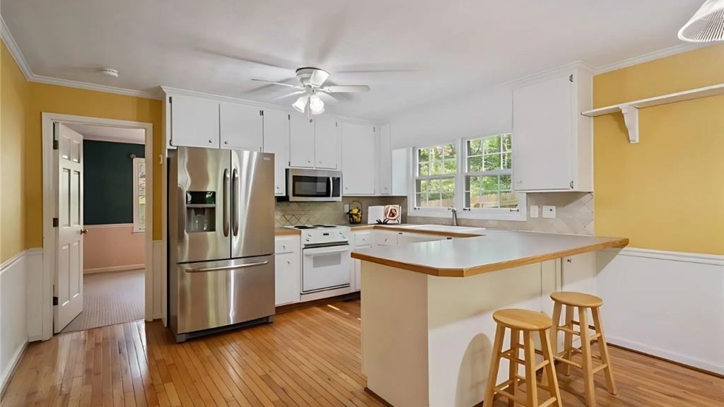 A bright kitchen with yellow walls, wooden floors, white cabinets, a stainless-steel fridge, and a central island with stools.