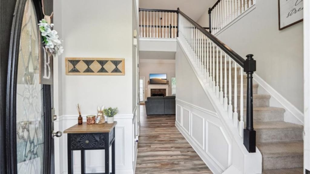 A bright foyer with a grand staircase, hardwood floors, a console table, and a view into a cozy living room.