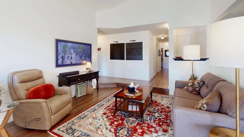 A cozy living room with a recliner, gray sofa, red rug, wooden coffee table, and artwork, lit by stylish floor lamps.