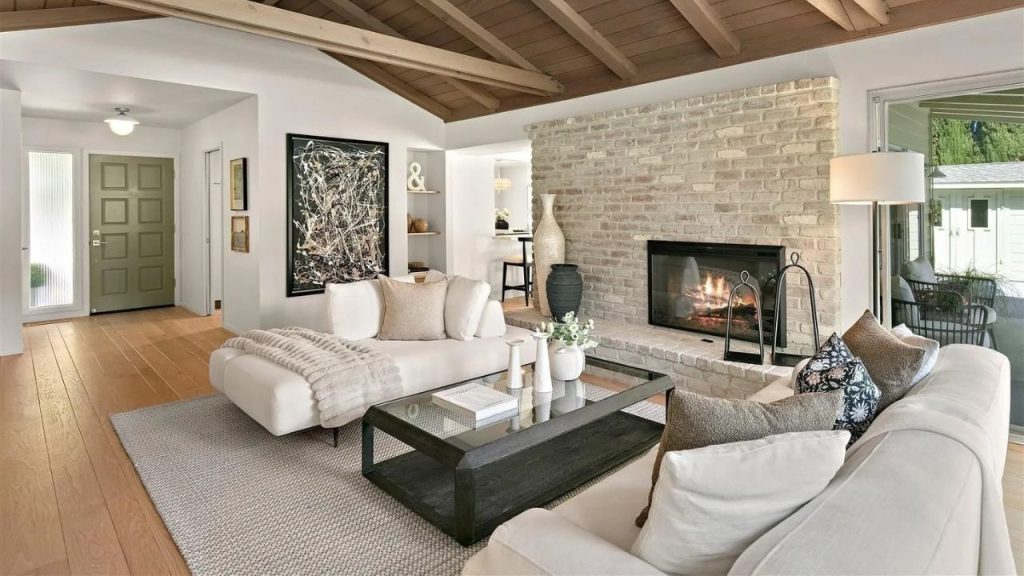 A cozy living room with a brick fireplace, plush sofa, and wooden beams, bathed in natural light from large windows.