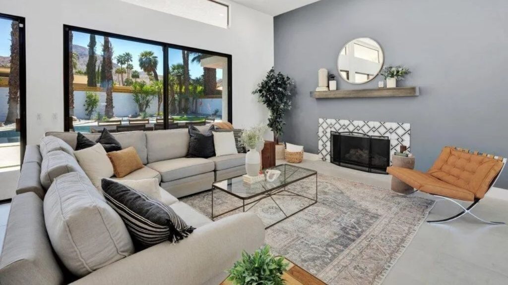 A cozy desert living room with a sectional sofa, fireplace, and large windows overlooking palm trees.
