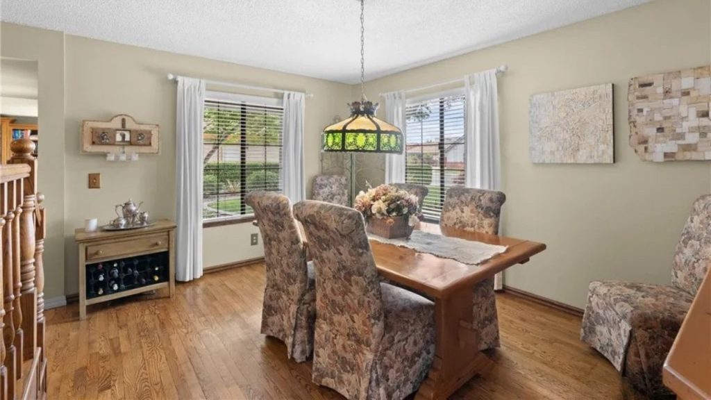 A cozy dining room with a wooden table, floral-patterned chairs, a stained-glass chandelier, and large windows letting in natural light.