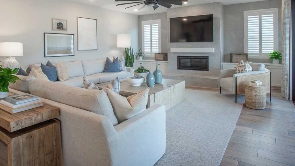 A bright living room with a light gray sectional sofa, blue pillows, a modern fireplace, a mounted TV, light wood floors, and large windows with white shutters.