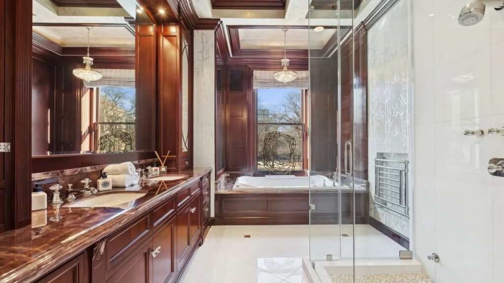 An opulent bathroom with a deep tub, double vanity in mahogany, a glass shower, and a chandelier, featuring large windows with a winter view.