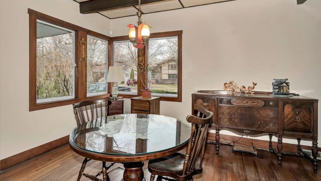 A vintage dining nook with a glass-top table, carved wooden chairs, an antique sideboard, a dual-lamp chandelier, and large windows framing a quiet neighborhood view.