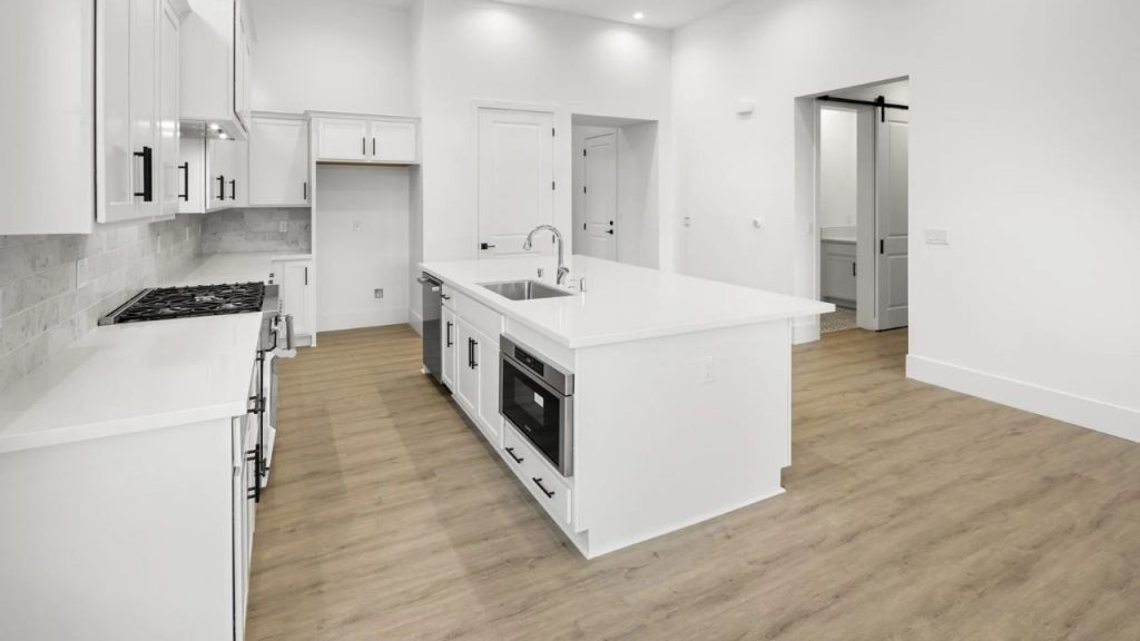 A modern kitchen with white cabinetry, a large island with a sink and oven, light wood floors, a subway tile backsplash, and a sliding barn door to a pantry.
