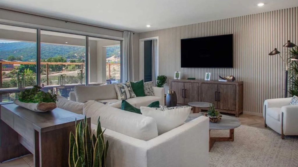 A cozy living room with a plush white sofa, green accent pillows, a wooden console, and large windows overlooking rolling hills.