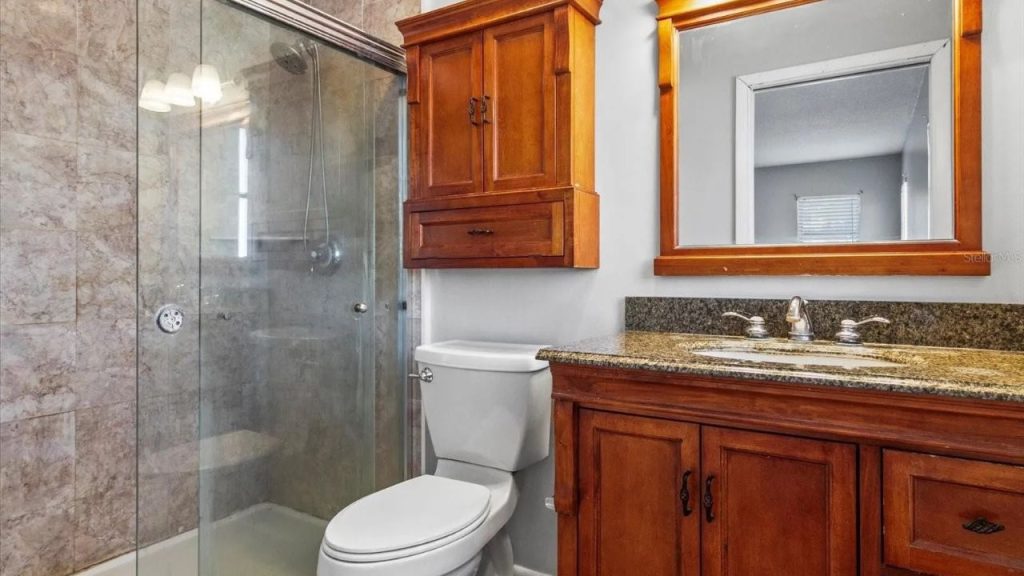 A stylish bathroom with wooden cabinets, a granite double sink, a large mirror, a glass shower with marble tiles, and soft lighting.