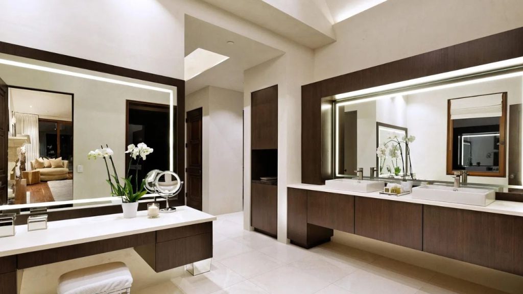 A stylish bathroom with dual dark wood vanities, backlit mirrors, white orchids, and marble floors, with a cozy room visible in the reflection.