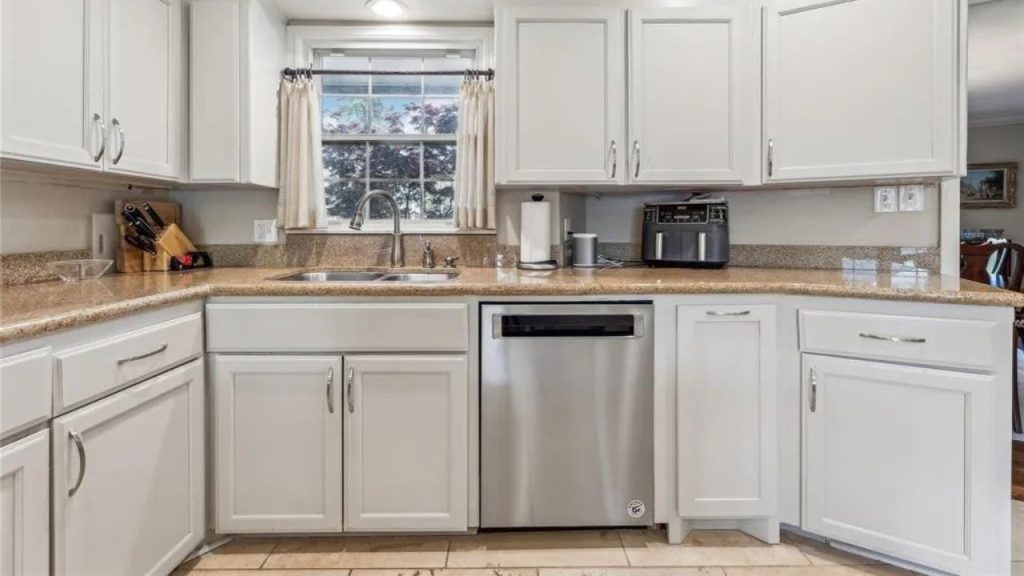 A bright kitchen with white cabinets, granite countertops, stainless-steel appliances, and a window with curtains.