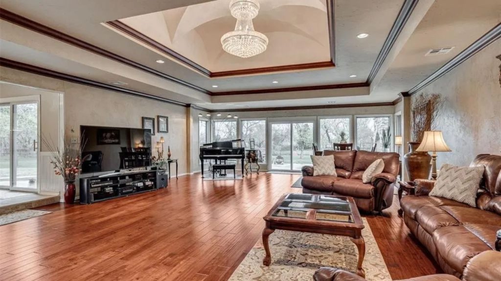 Spacious living room with leather sofas, a chandelier, piano, and large windows, featuring warm hardwood floors.