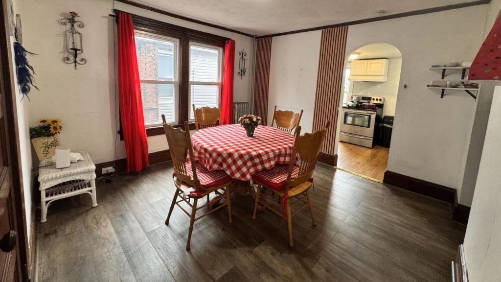 A cozy dining area with a checkered tablecloth, wooden chairs, and large windows with red curtains.