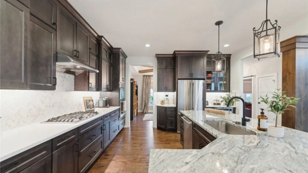 A modern kitchen with dark cabinets, marble countertops, and pendant lights near an open dining area.